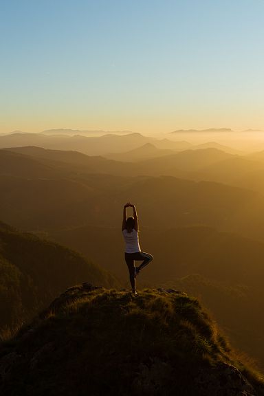 Person steht in Yoga-Position auf einem Berggipfel bei Sonnenuntergang, umgeben von hügeliger Landschaft und goldenem Licht des Abendhimmels.