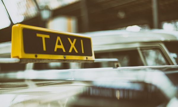 Nahaufnahme eines Taxis mit gelbem "Taxi"-Schild auf dem Dach, unscharfer Hintergrund von weiteren Fahrzeugen sichtbar, städtische Umgebung.
