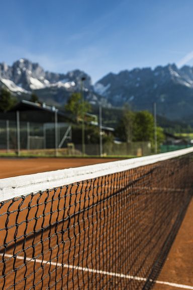 A clay tennis court with a net in the foreground and the Wilder Kaiser mountains in the background under a blue sky.
