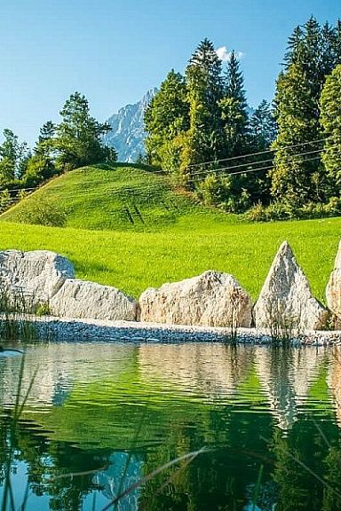 A serene landscape featuring a pond with clear reflections, surrounded by green hills and pine trees, against the backdrop of the Wilder Kaiser mountains.