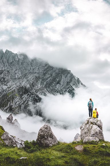 Zwei Wanderer stehen auf einem Felsen in den Alpen, umgeben von grünem Gras und Nebel, mit einem imposanten Bergmassiv im Hintergrund.