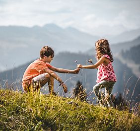 Zwei Kinder spielen auf einer Wiese in den Bergen. Der Junge kniet, während das Mädchen Blumen hält. Im Hintergrund sind nebelige Berge zu sehen.