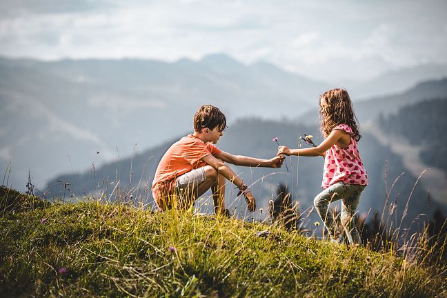 Zwei Kinder spielen auf einer Wiese in den Bergen. Der Junge kniet, während das Mädchen Blumen hält. Im Hintergrund sind nebelige Berge zu sehen.