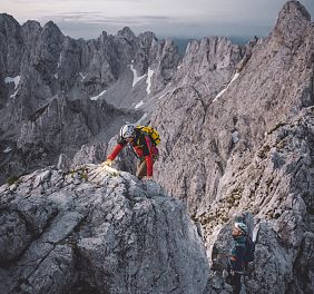 Two climbers in safety gear navigate a steep, rocky terrain on a mountain. The backdrop is rugged with gray rock formations and patches of snow.