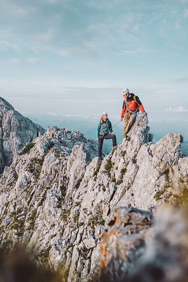 Two hikers in bright clothing climb a rugged, rocky mountain ridge with dramatic peaks in the background under a partly cloudy sky.