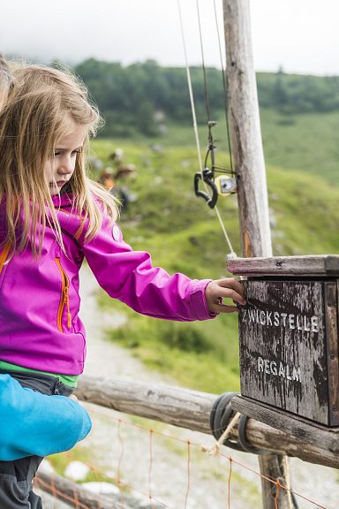 Eine Frau hält ein Kind, das neugierig eine hölzerne Schilderbox im Grünen betrachtet. Beide tragen bunte Outdoorjacken. Im Hintergrund sind Berge zu sehen.