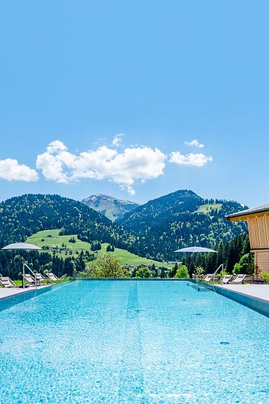 An outdoor pool with wooden lodges and sun umbrellas, set against the backdrop of the Wilder Kaiser mountains under a bright blue sky.