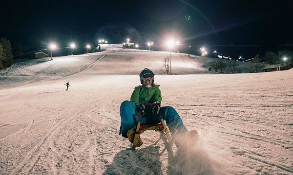 Person beim Nachtrodeln auf einer beleuchteten Skipiste, umgeben von Schneelandschaft. Im Hintergrund sind weitere Skisportler zu sehen.