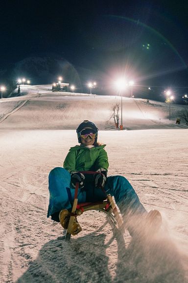 Person beim Nachtrodeln auf einer beleuchteten Skipiste, umgeben von Schneelandschaft. Im Hintergrund sind weitere Skisportler zu sehen.