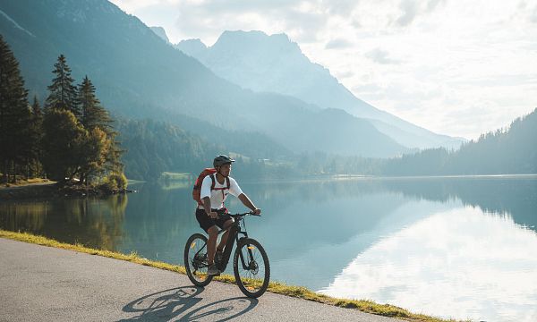Ein Radfahrer auf einem Radweg neben einem ruhigen See in einer Berglandschaft. Die Sonne scheint, und die Berge spiegeln sich im Wasser wider.