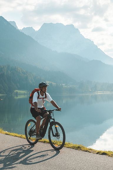 Ein Radfahrer auf einem Radweg neben einem ruhigen See in einer Berglandschaft. Die Sonne scheint, und die Berge spiegeln sich im Wasser wider.