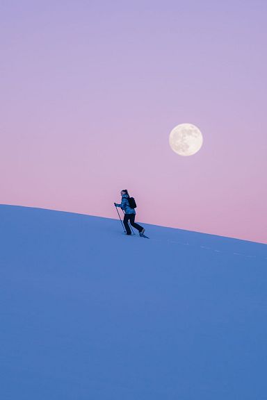 Eine Person wandert bei Sonnenuntergang in einer verschneiten Landschaft, der Vollmond steht am Himmel. Der Horizont zeigt zarte Blau- und Lilatöne.
