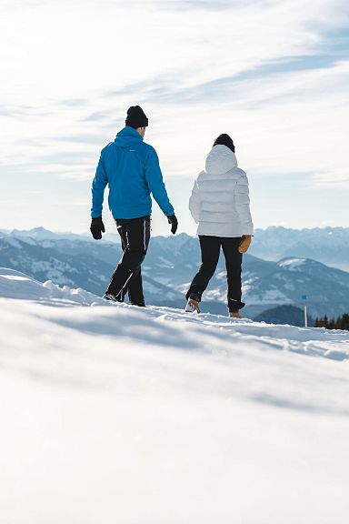 Zwei Personen spazieren im Winter auf einem schneebedeckten Bergpfad, mit einem Gipfelkreuz und einer weiten Berglandschaft im Hintergrund.
