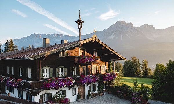 Traditionelles Tiroler Holzhaus mit blühenden Geranien, im Hintergrund das beeindruckende Bergmassiv des Wilden Kaisers bei Sonnenaufgang.