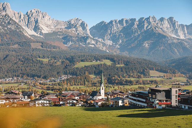 Panoramablick auf eine Tiroler Ortschaft vor einem imposanten Bergmassiv bei klarem Himmel, umgeben von grünen Wiesen und dichten Wäldern.