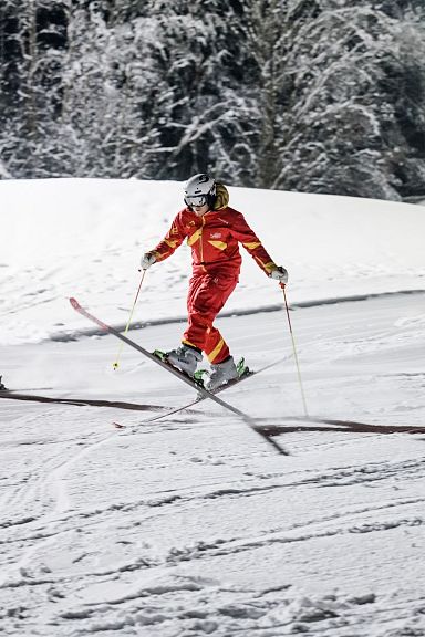 Drei Skifahrer in roten Anzügen fahren synchron eine Piste hinunter, umgeben von Schneebäumen. Es ist Nacht, und die Szene ist von Flutlicht beleuchtet.