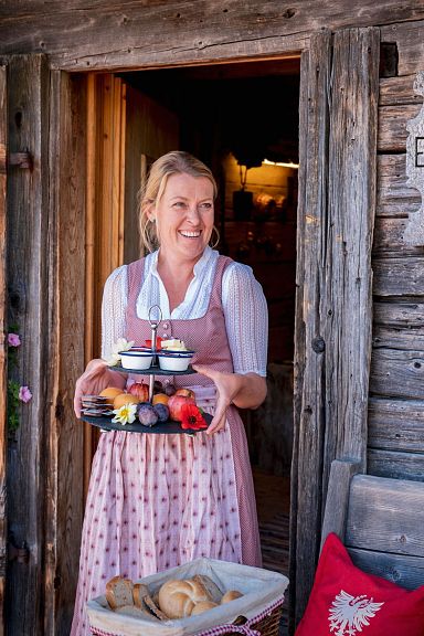 Eine Frau in traditioneller Tracht steht vor einer Holzhütte und hält ein Tablett mit Gebäck. Neben ihr ist eine Bank mit Tiroler Dekoration.