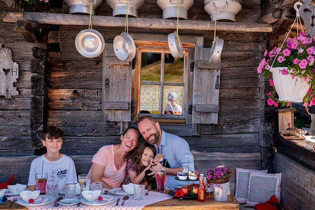 A family enjoying breakfast at a rustic wooden table outside a log cabin, surrounded by hanging flowers and traditional milk churns.