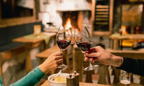 Two people clinking wine glasses in a cozy, rustic setting with a warm fireplace in the background.