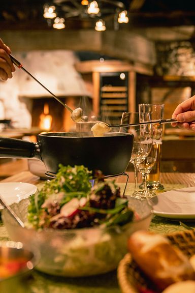 Zwei Personen genießen Fondue in einer gemütlichen Alpenhütte. Auf dem Tisch stehen Brot, Salat und Dips. Eine warme, einladende Atmosphäre herrscht vor.