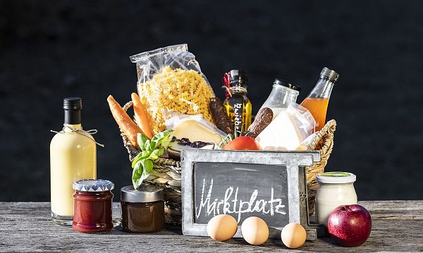 A rustic wooden table displays a basket with pasta, eggs, a chalkboard sign, a bottle of juice, and various jars, reflecting a farmers market setup.