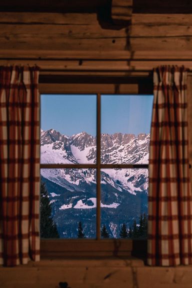 Cozy wooden cabin interior with checkered curtains, window view of snow-covered Wilder Kaiser mountains. Lantern on wall adds to rustic charm.