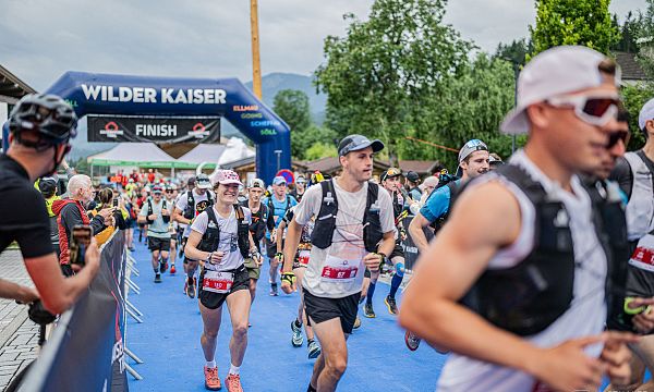 Runners in athletic gear approaching a finish line at a Wilder Kaiser event, surrounded by greenery and mountains in the background.