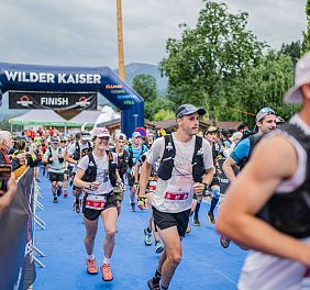 Runners in athletic gear approaching a finish line at a Wilder Kaiser event, surrounded by greenery and mountains in the background.