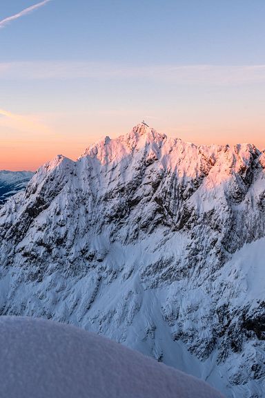 Snow-covered peaks of the Wilder Kaiser at sunrise, glowing with warm colors against a clear sky. The light highlights the rugged mountain landscape.