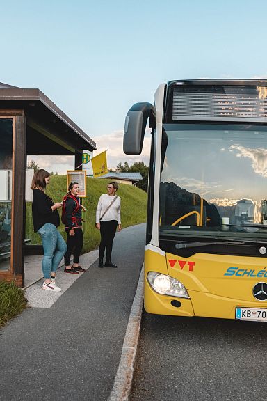 Ein gelber Linienbus hält an einer Haltestelle auf einer Landstraße. Mehrere Personen warten neben einem überdachten Wartebereich. Die Sonne geht unter.