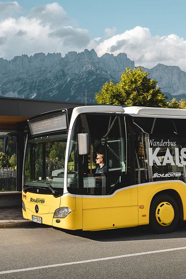 A yellow bus at a stop with people walking nearby, set against the backdrop of the Wilder Kaiser mountains and a partly cloudy sky.