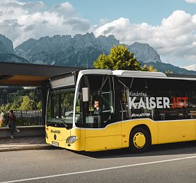 Ein gelber Linienbus steht an einer Haltestelle vor einer Bergkulisse mit dem Wilder Kaiser im Hintergrund. Zwei Personen gehen am Gehweg vorbei.