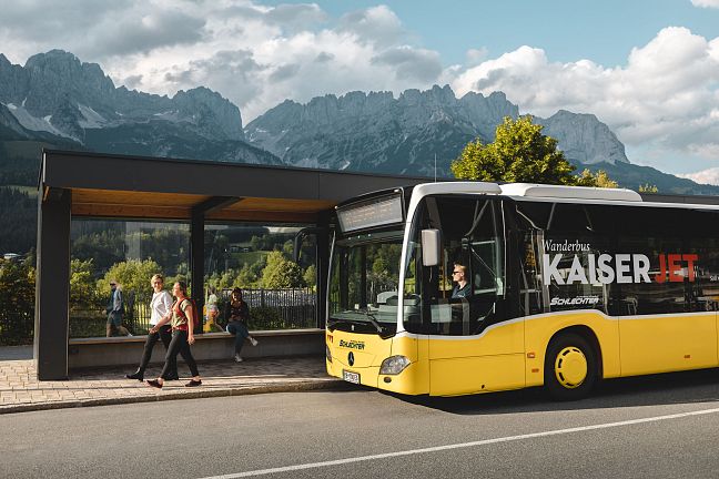 Ein gelber Linienbus steht an einer Haltestelle vor einer Bergkulisse mit dem Wilder Kaiser im Hintergrund. Zwei Personen gehen am Gehweg vorbei.