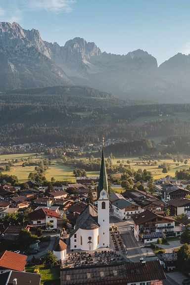 A charming village with a prominent church spire lies under the impressive Wilder Kaiser mountain range, illuminated by the gentle morning light.