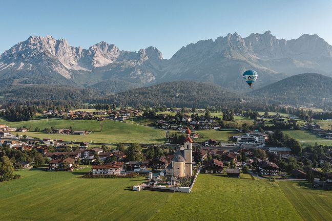 Landschaft mit grünen Wiesen, einem Dorf und dem imposanten Bergmassiv des Wilden Kaisers im Hintergrund. Ein Heißluftballon schwebt am klaren Himmel.