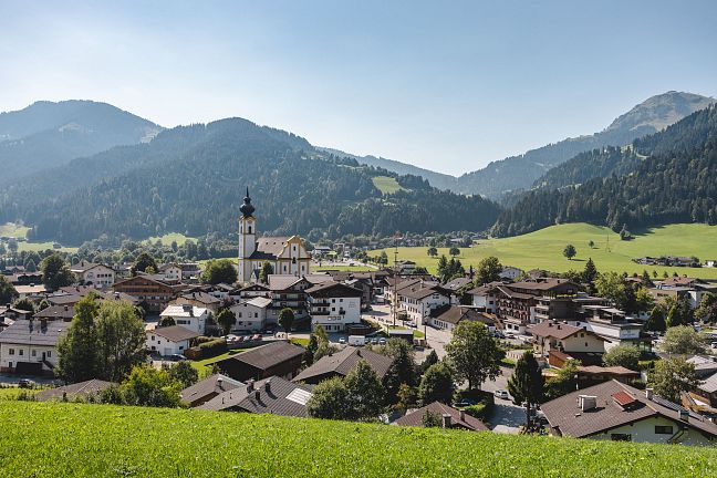 Österreichisches Dorf mit Kirche im Zentrum, umgeben von grünen Hügeln und Wäldern, bei sonnigem Wetter. Traditionelle Tiroler Architektur dominiert die Szene.