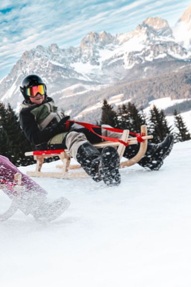 Zwei Personen rodeln fröhlich einen verschneiten Hang hinunter, umgeben von Nadelbäumen und einem beeindruckenden Bergmassiv im Hintergrund.