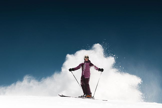 Skier in purple outfit on fresh snow, clear blue sky, Wilder Kaiser mountains in the background, showcasing a thrilling winter scene.