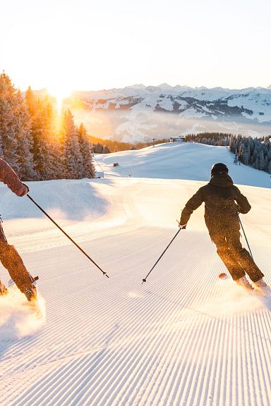 Zwei Skifahrer fahren bei Sonnenaufgang eine schneebedeckte Piste hinunter, umgeben von verschneiten Bäumen und Bergen im Hintergrund.