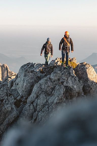 Zwei Wanderer stehen auf einem felsigen Berggipfel mit beeindruckender Aussicht auf umliegende Täler und Berge bei Sonnenuntergang.