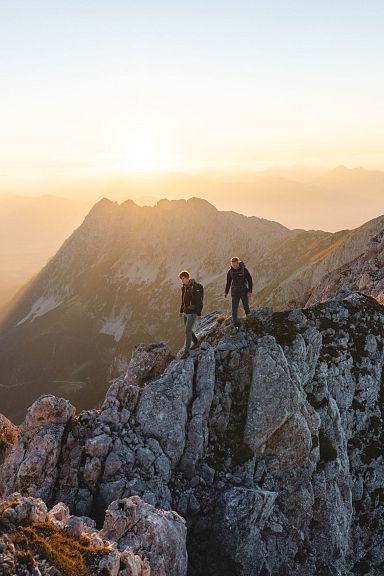 Zwei Wanderer stehen bei Sonnenaufgang auf einem felsigen Berggipfel. Im Hintergrund erheben sich weitere Berge und ein See ist in der Ferne sichtbar.