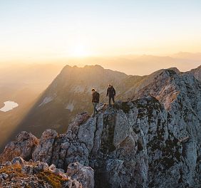 Zwei Wanderer stehen bei Sonnenaufgang auf einem felsigen Berggipfel. Im Hintergrund erheben sich weitere Berge und ein See ist in der Ferne sichtbar.