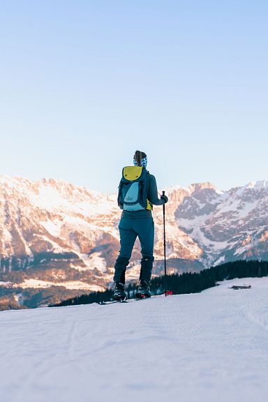 A person in winter gear with a backpack and ski poles stands on a snowy slope, admiring the Wilder Kaiser mountains under a clear blue sky at sunset.