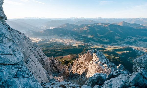 Blick von einem felsigen Berggipfel auf ein weites Tal mit sanften Hügeln und Wäldern, unter einem klaren blauen Himmel in den Alpen.