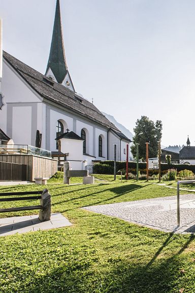 Ein Dorfplatz bei Sonnenaufgang mit einer Kirche, einem Baum und einem kleinen runden Brunnen. Im Hintergrund sind Häuser und ein bewaldeter Hügel zu sehen.