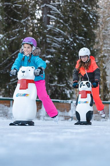 Zwei Kinder im Schnee mit Eislaufhilfen in Form von Tieren, umgeben von schneebedeckten Bäumen. Beide tragen bunte Winterkleidung und Helme.