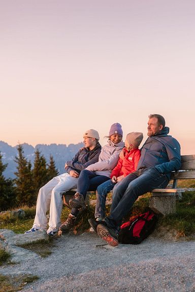 A family sitting on a bench, enjoying the sunset at Wilder Kaiser with mountains in the background, under a clear sky. The scene is calm and picturesque.