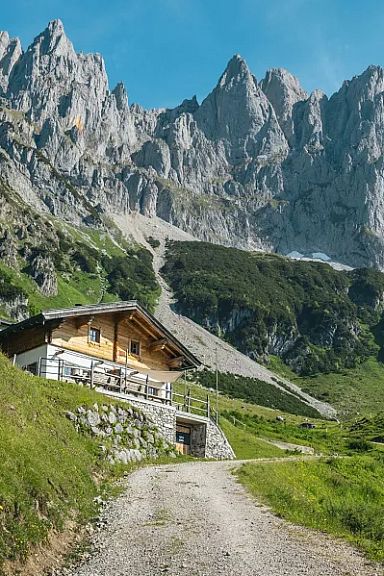 Berglandschaft mit grünen Wiesen und einem holzverkleideten Haus im Vordergrund, dahinter ragen schroffe, felsige Gipfel unter einem blauen Himmel empor.