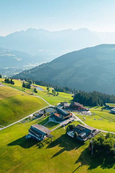 Aerial view of green hills and scattered houses beneath the towering Wilder Kaiser mountains in Tirol, Austria, on a clear day.