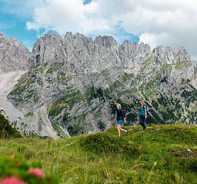 Zwei Wanderer spazieren auf einer grünen Wiese vor einem imposanten Bergmassiv in den Alpen. Der Himmel ist bewölkt, mit blauen Flecken sichtbar.